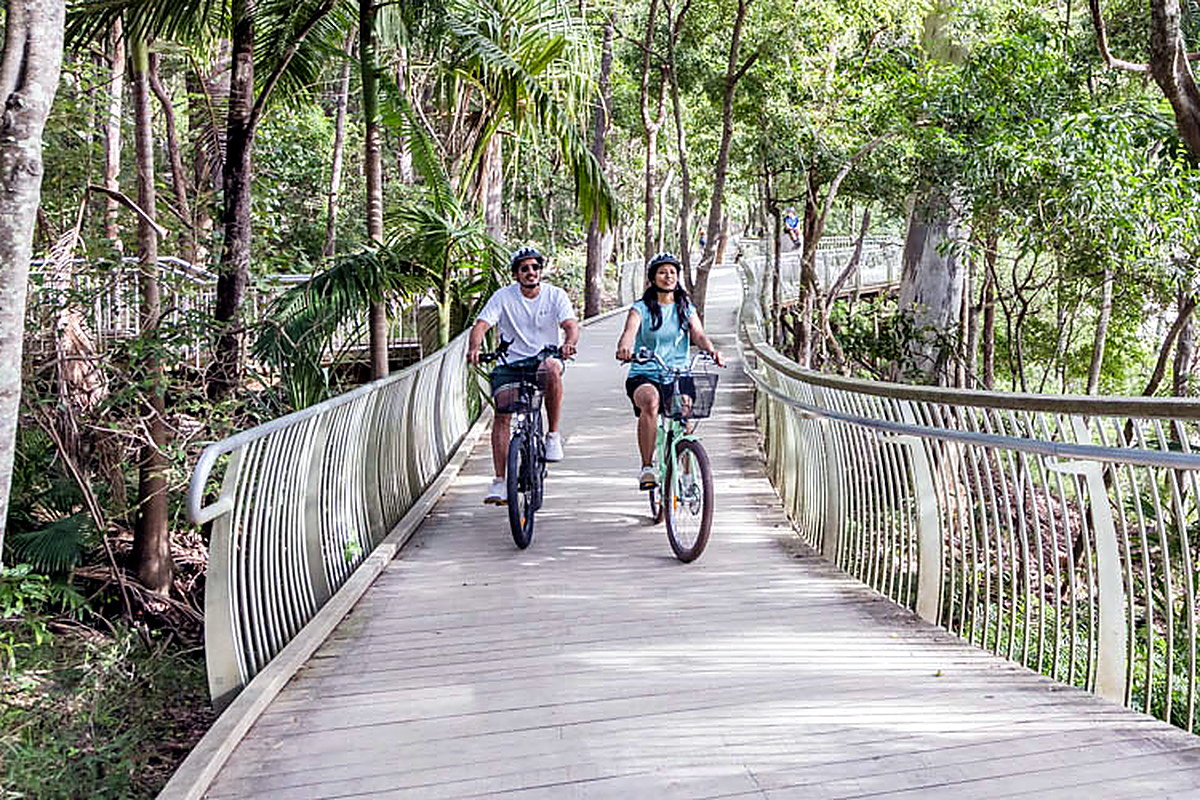 Two riders on a timber boardwalk through palms and trees in Noosa
