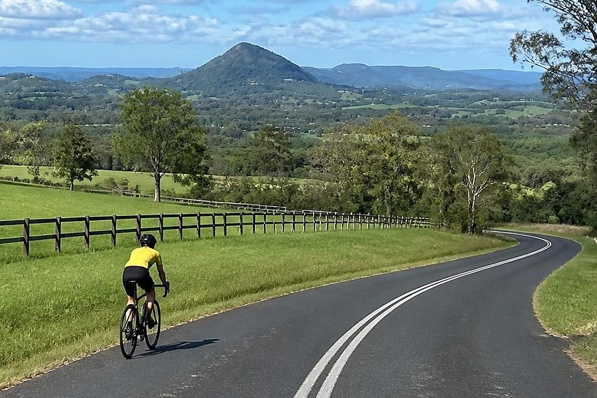 Road cyclist on a winding sealed hinterland road past green paddocks and mountain views, Sunrise Hills Loop road ride from Bike On Noosa